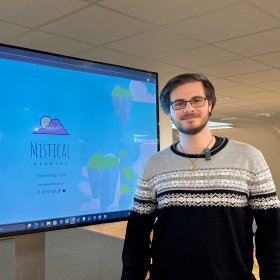 Portrett av spilldesigner på kontor Mann med kort, mørkt hår og briller står foran en TV-skjerm på et kontor. Han har på seg en langermet genser i svart og grått. I bakgrunnen skimtes et felles kontorområde.