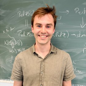Portrett av Matematiker. En mann med brunt, kort hår og grønn, kortermet t-skjorte står foran en grønn tavle. Bak ham er det skrevet matematiske formler. Han ser mot kamera og smiler.