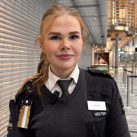 Kvinne med langt oransjegult hår i strikk smiler til kamera. Hun har på seg hvit skjorte, slips og mørkegrå genser som del av vekteruniform. I bakgrunnen skimtes deler av arbeidsplassen på Gardermoen.