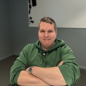 En mann sitter ved et bord med armene krysset foran seg og ser mot kamera med et svakt smil. Han har kort, gråsprengt hår og er kledd i en grønn hettegenser. På håndleddet har han en klokke med metallrem. Bak ham er en grå vegg med et enkelt kunstverk i hvitt og svart.