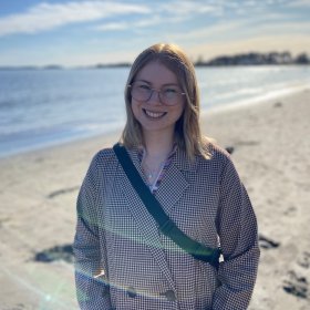 En kvinne smiler mot kameraet på en solfylt strand. Hun har briller, skulderlangt blondt hår og rutete frakk. Bak henne skimtes sjøen, himmelen og solskinnet.