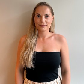 En dame med langt, blondt hår står inntil en hvit vegg og smiler med munnen lukket. Hun har på seg en svart tubetopp og et beige skjørt.