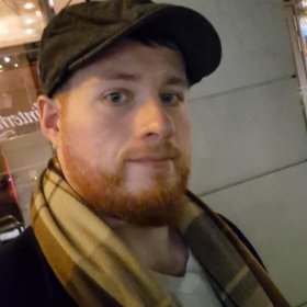 En mann med rødt skjegg tar et nærbilde av seg selv utendørs i mørket. Han har på seg en mørk caps, et rutete skjerf og en mørk jakke. I bakgrunnen er det en bygning med opplyste vinduer.