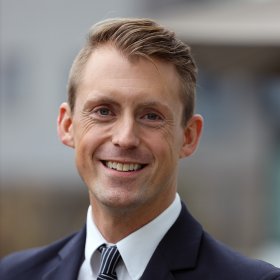 En mann i uniform står ute og smiler varmt mot kamera. Han har kort, blondt hår gredd til siden og lyseblå øyne. Han bærer en mørk marineblå kabinpersonale-uniform med hvit skjorte og stripete slips i rødt, hvitt og blått. På jakkeslaget sitter et lite rødt merke og et vingemerke med Norwegian-logoen.