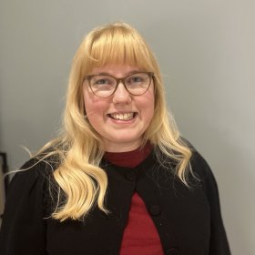 En jente med blondt hår og briller smiler til kamera. Hun står foran en grå vegg og har på seg en mørk cardigan med rød t-skjorte under.