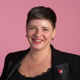 En kvinne med kort, mørkt hår smiler mot kamera. Hun har på seg en svart blazer med en rød knapp på jakkeslaget, gulløredobber og en sølvkjede. Bakgrunnen er ensfarget rosa.