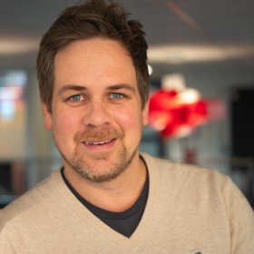 Et nærportrett av en mann som lener seg lett mot en hvit vegg. Han smiler svakt, og bakgrunnen består av et kontormiljø med røde lamper som gir varme farger.