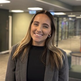 En dame med langt brunt hår. Damen har på seg en svart genser og en grå blazer. Hun står i et åpent rom med grønne vegger. Hun smiler med tennene. 