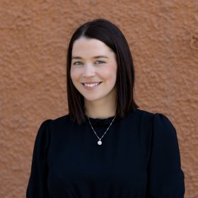 Ei dame med mørkt hår. Hun har svart kjole og et sølvsmykke på seg. Hun ser rett i kameraet og smiler. Hun står foran en brun vegg.