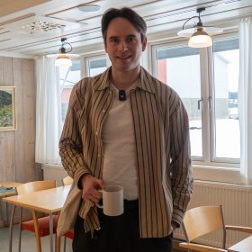 En mann står i et personalrom med lys trepanel og store vinduer. Han holder en kaffekopp i høyre hånd og ser mot kamera. Rundt ham står bord og stoler, og rommet har en rolig og uformell atmosfære.