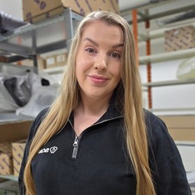 En dame med langt blondt hår smiler, hun har på seg en svart genser med glidelås som er litt åpen. Bak henne er det et stativ med ulike esker.
