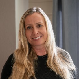 En kvinne med langt, lyst hår smiler mot kamera. Hun har på seg en sort genser og står innendørs med nøytrale gardiner og vegger i bakgrunnen.