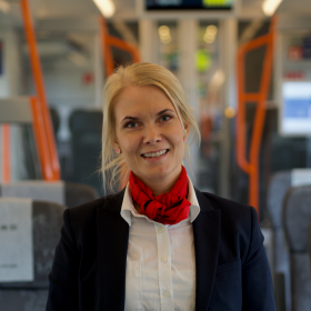 En kvinne i blå uniform med hvit skjorte og rødt skjerf smiler til kamera. I bakgrunnen er det togseter og informasjonsskjermer.