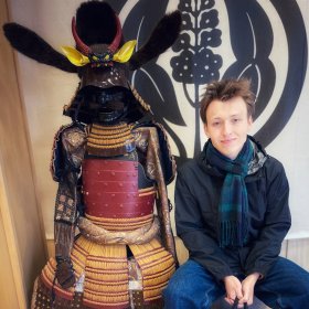 Marius Teig-Hermanrud En ung mann sitter ved siden av en tradisjonell japansk samuraidraktsfigur. Han smiler. Mannen i et museum eller kulturhus.
