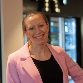 En kvinne smiler til kamera. Hun har på en rosa blazer, og står i et rom med et stor kjøleskap med glassdører i bakgrunn. 