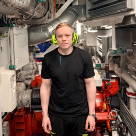 En mann står i et trangt maskinrom omgitt av store motorer, rør, ventilasjonssystemer og teknisk utstyr. Han har på seg hørselsvern og arbeidsklær, og ser rett mot kameraet. Rommet er tettpakket med maskineri og metallrør i ulike størrelser.