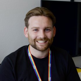Mann med svart t-skjorte og brunt hår. Rund halsen har han en et nøkkelknippe med regnbuefarger. Mannen smiler til kamera.