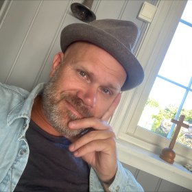 Portrett av en mann med hatt og skjegg i svart t-skjorte og dongerijakke