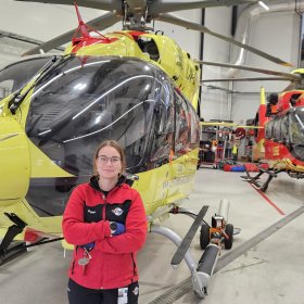 En kvinne med briller og rød arbeidsjakke står med armene i kors foran et stort, gult helikopter i en lys hangar. Et mindre rødt og gult helikopter og et svart og rødt kjøretøy står lenger bak.