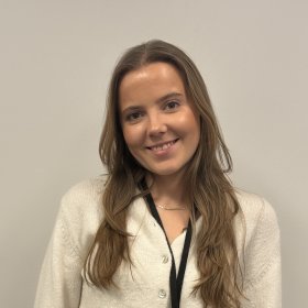 En kvinne med langt brunt hår og med hvit jakke står foran en lys vegg