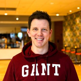 En mann med kort, mørkt hår smiler mot kamera. Han har på seg en mørkerød hettegenser med teksten «GANT» på brystet. Bildet er tatt innendørs i et lyst rom med varm belysning og uskarp bakgrunn.