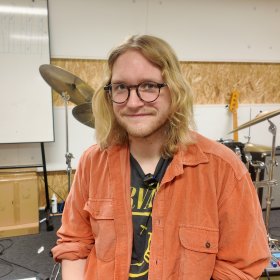 En person sitter i et musikkrom og ser inn i kameraet. Han har skulderlangt blondt hår, briller og har på seg en åpen skjorte med en band-t-skjorte under. Bak han står et trommesett, en forsterker, gitarer og andre instrumenter. Veggene er dekket av sponplater, og et whiteboard står til venstre i bildet.