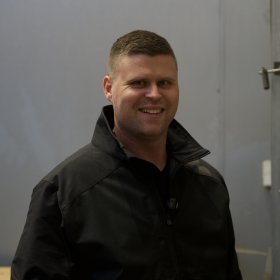 En mann smiler mot kameraet. Han står i et rom med stålvegger og har på seg en svart jakke med høy krage. På veggen til høyre henger en stålkonstruksjon