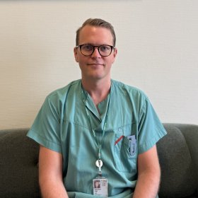 En mann i grønne sykehusklær sitter på en sofa og ser mot kamera. Han har korte, lyse hår, briller og ID-kort rundt halsen. Bak ham er en lys vegg med et innrammet bilde.