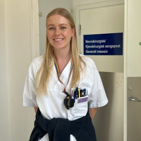 En ung kvinne i hvite sykehusklær står i en korridor på sykehuset og smiler mot kamera. Hun har langt, blondt hår og navneskilt festet på brystet. Bak henne er en dør med skilt der det står «Nevrokirurgisk/Kjevekirurgisk sengepost Generell intensiv».