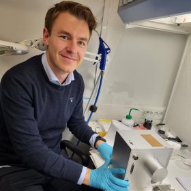 En mann sitter ved en labbenk med blå engangshansker og holder et sølvfarget optisk instrument. Rundt ham ligger presisjonsdeler og laboratorieutstyr, og han smiler mot kameraet.