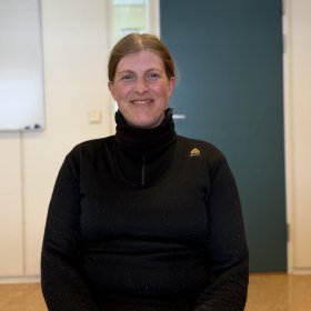 En kvinne smiler mot kamera. Hun har på seg en svart genser med høy hals og har håret trukket bakover. I bakgrunnen ser man et enkelt rom med en grønn dør og en tavle på veggen.