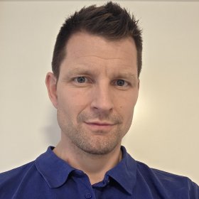 En mannlig behandler i blå poloskjorte står foran en lys, ensfarget vegg. Han ser rett mot kamera med et rolig og profesjonelt uttrykk. Han har kort, mørkt hår og lett skjeggstubb.