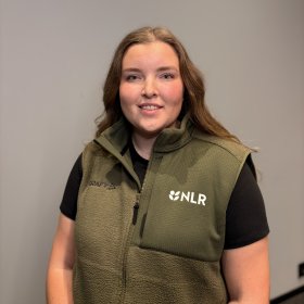 Dame med brunt hår og grønn vest over en svart t-skjorte smiler til kamera