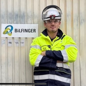 Plastmekaniker Simen Lauritzen En mann står i gul og svart arbeidsdress og lener seg mot en industrivegg. Han har på seg vernebriller, og på hodet har han en hjelm. Til venstre for han er et skilt der det står "Bilfinger".