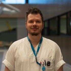 En mann står innendørs i et større bygg med åpent rom og belysning i taket. Han har på seg hvite arbeidsklær og et nøkkelkort i snor rundt halsen. Bak ham er det gangarealer og åpne soner.