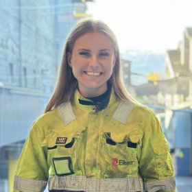 Dame med lyst hår og gule arbeidsklær smiler mot kamera.
