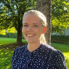 En kvinne i blå skjorte og lyst hår i hestehale står i en park med grønn plen og store trær