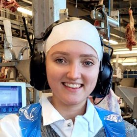 En dame i hvitt arbeidstøy og et blått plastikkforkle. På hodet har hun en hvit lue og hørselsvern. Hun står i et produksjonslokale. Fra taket henger det slakt.