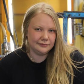 Bilde av en dame i sorte klær som sitter i det som ser ut som et verksted. Hun har blondt hår. 