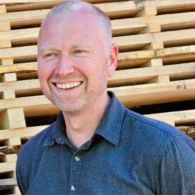En smilende mann står utendørs foran stablede treplanker. Han har kort, lyst hår, skjeggstubb og mørk skjorte, og ser litt til siden med avslappet uttrykk.