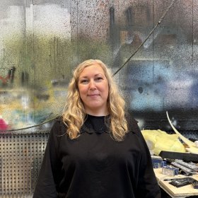En kvinne med langt, krøllete blondt hår smiler mot kameraet. Hun har på seg en svart genser. Bak henne er en vegg full av ulike farger.