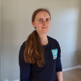 En kvinne med langt, brunt hår og hestehale står inne i et lyst rom. Hun har på seg en mørk blå genser med firmalogo og ser rett mot kamera. I bakgrunnen står et lite bord og et skap.