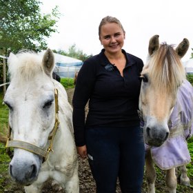 Ei dame i mørke klær. Hun står mellom to små hester. Hun er ute. 