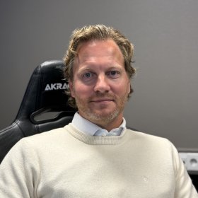 En mann med lyst, kort, bølgete hår og skjegg sitter i en svart kontorstol og ser mot kameraet. Han har på seg en lys skjorte under en hvit genser.