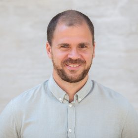 En mann i midten av 30-årene står foran en lys, nøytral bakgrunn og ser rett inn i kamera. Han har kort mørkt hår, kort skjegg og et vennlig smil. Han har på seg en lys, knappet skjorte med krage. Bildet er et klassisk portrettbilde med rolig bakgrunn og naturlig lys.