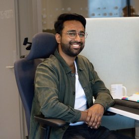 En mann i mørkegrønn skjorte, hvit t-skjorte og briller. Han smiller og ser rett i kamera. Han er på kontoret.