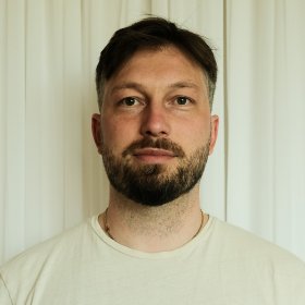 En mann med mørkt hår og skjegg og lys t-skjorte står foran en lys vegg
