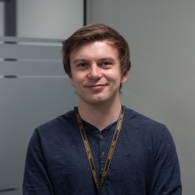 En person med ID-bånd rundt halsen står innendørs foran en vegg med matterte glasspaneler. Personen ser mot kamera og smiler svakt.