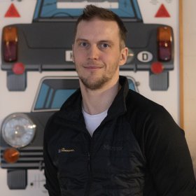 En mann i svart jakke og hvit t-skjorte. Bak han er det bilder av gamle bilder. Han er inne.