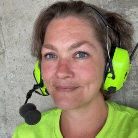 En dame med mørkt hår og gule øreklokker står foran en grå vegg.